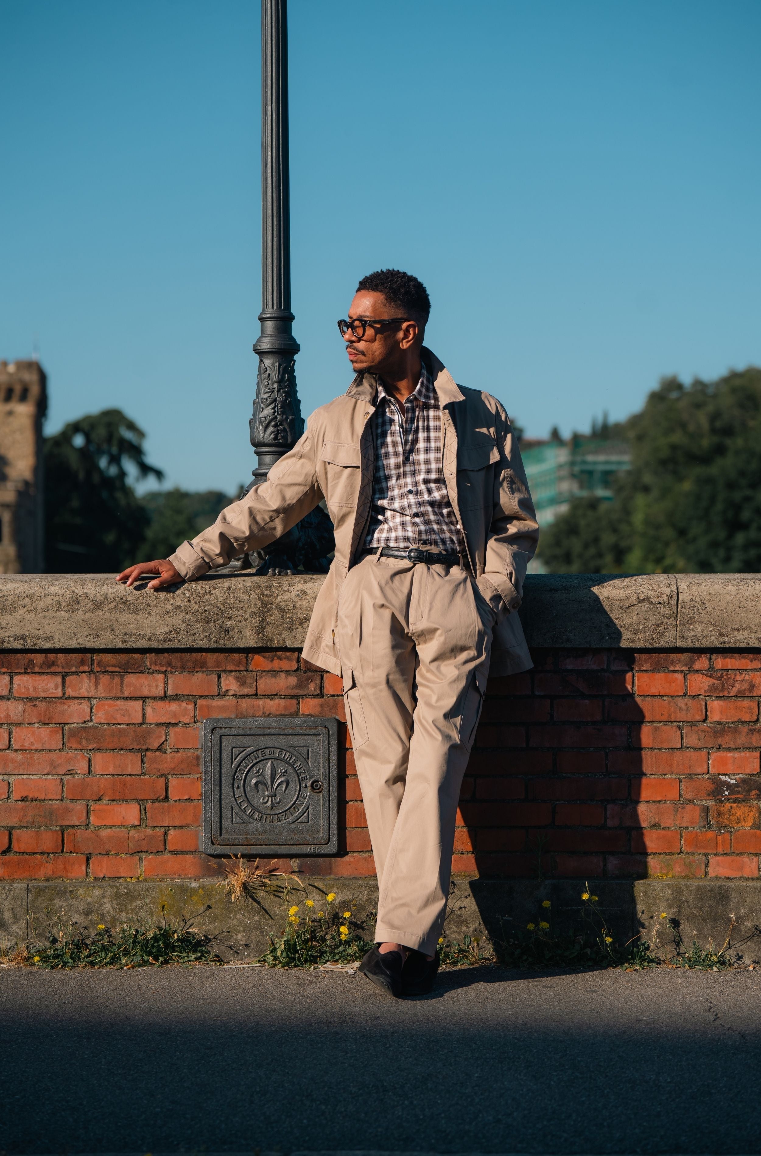 A man wearing a beige trench coat and matching trousers, a plaid shirt, and sunglasses, leaning against a brick wall outdoors under a clear blue sky.