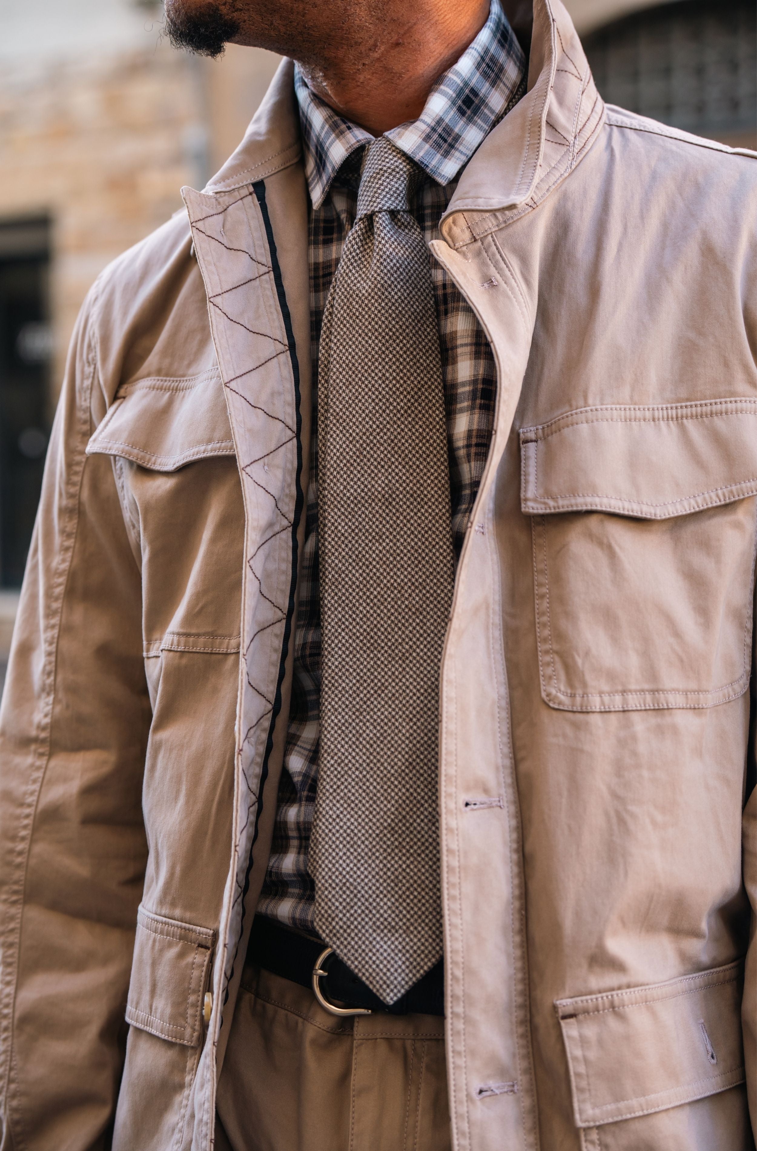 A close-up of a man wearing a beige utility jacket over a plaid shirt and a textured gray tie, with tan trousers and a black belt.