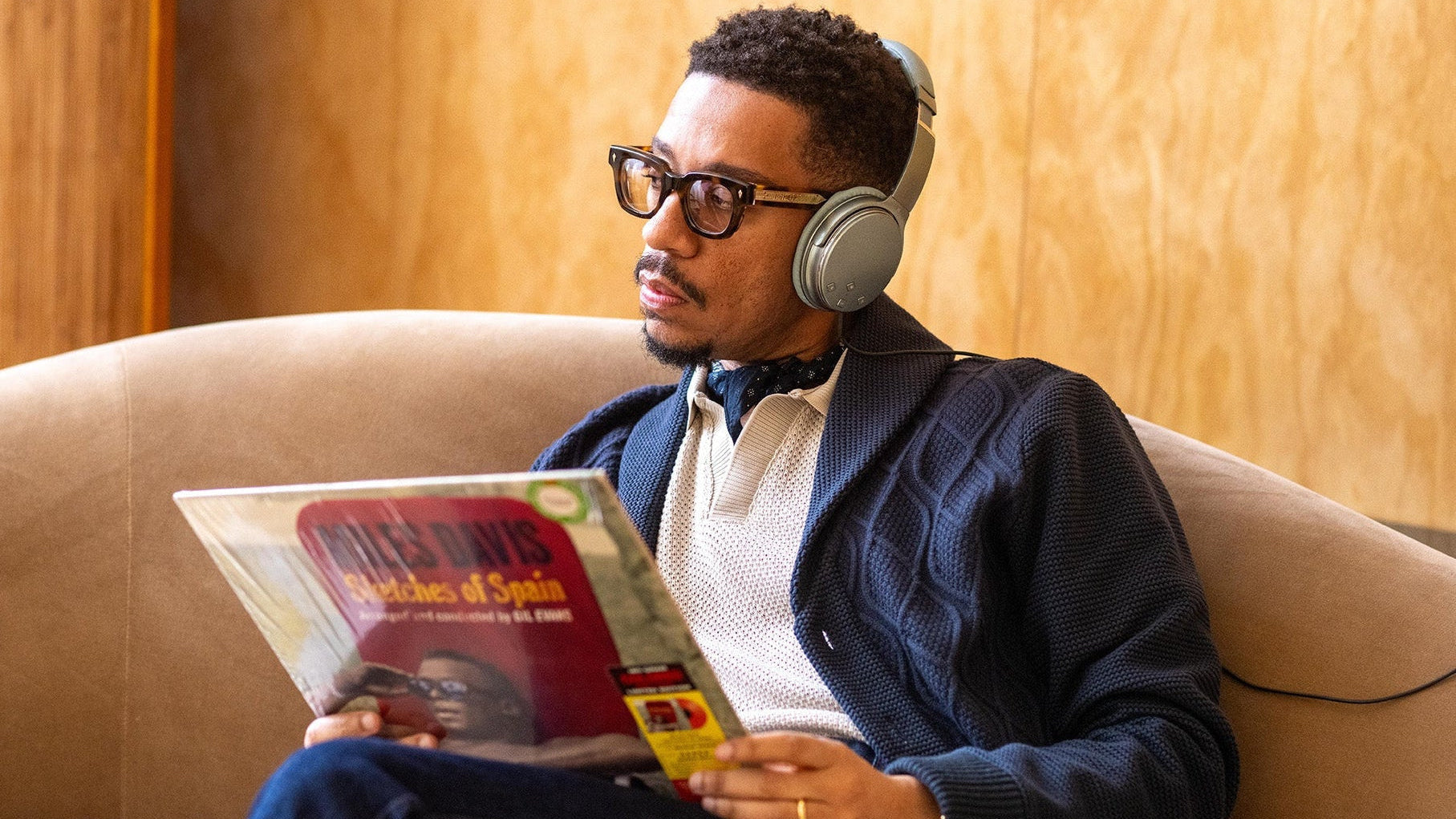 A man wearing over-ear headphones and glasses sits on a beige couch, reading a vinyl record sleeve titled 'Poulis Davis Sketches of Spain'. He is dressed in a navy cable-knit cardigan over a cream knit sweater.